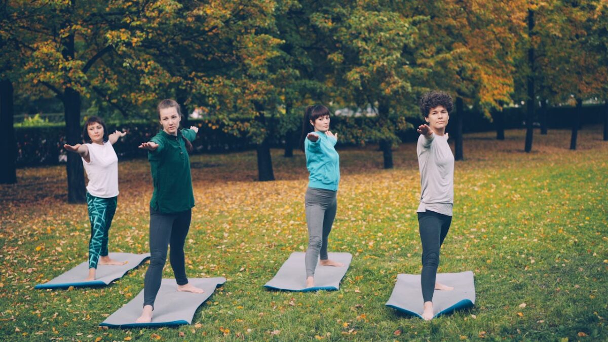 Four adults practicing yoga on mats outdoors in a scenic autumn park setting.